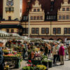 Blumenmarkt in der historischen Altstadt mit Verkaufständen unter grün-weißen Marktschirmen, Menschen beim Einkauf, im Hintergrund das prächtige Rathaus im gotischen Stil, lebendiges Stadtbild, Touristen und Einheimische.