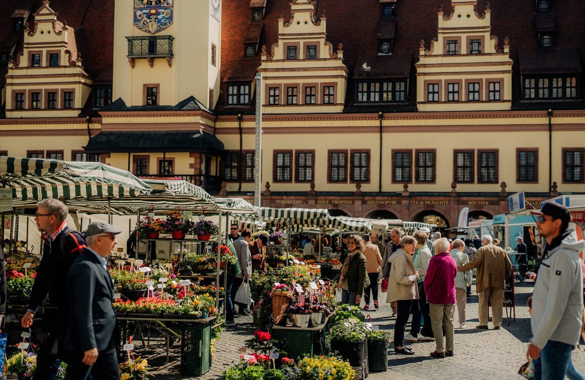 Blumenmarkt in der historischen Altstadt mit Verkaufständen unter grün-weißen Marktschirmen, Menschen beim Einkauf, im Hintergrund das prächtige Rathaus im gotischen Stil, lebendiges Stadtbild, Touristen und Einheimische.