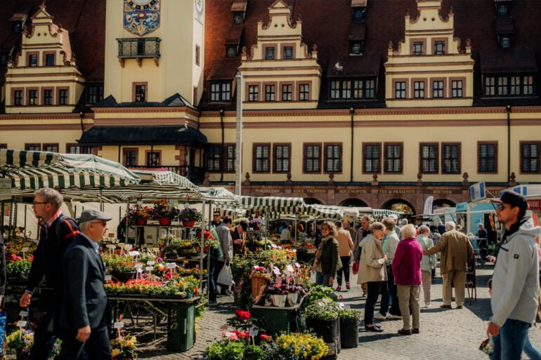 Blumenmarkt in der historischen Altstadt mit Verkaufständen unter grün-weißen Marktschirmen, Menschen beim Einkauf, im Hintergrund das prächtige Rathaus im gotischen Stil, lebendiges Stadtbild, Touristen und Einheimische.